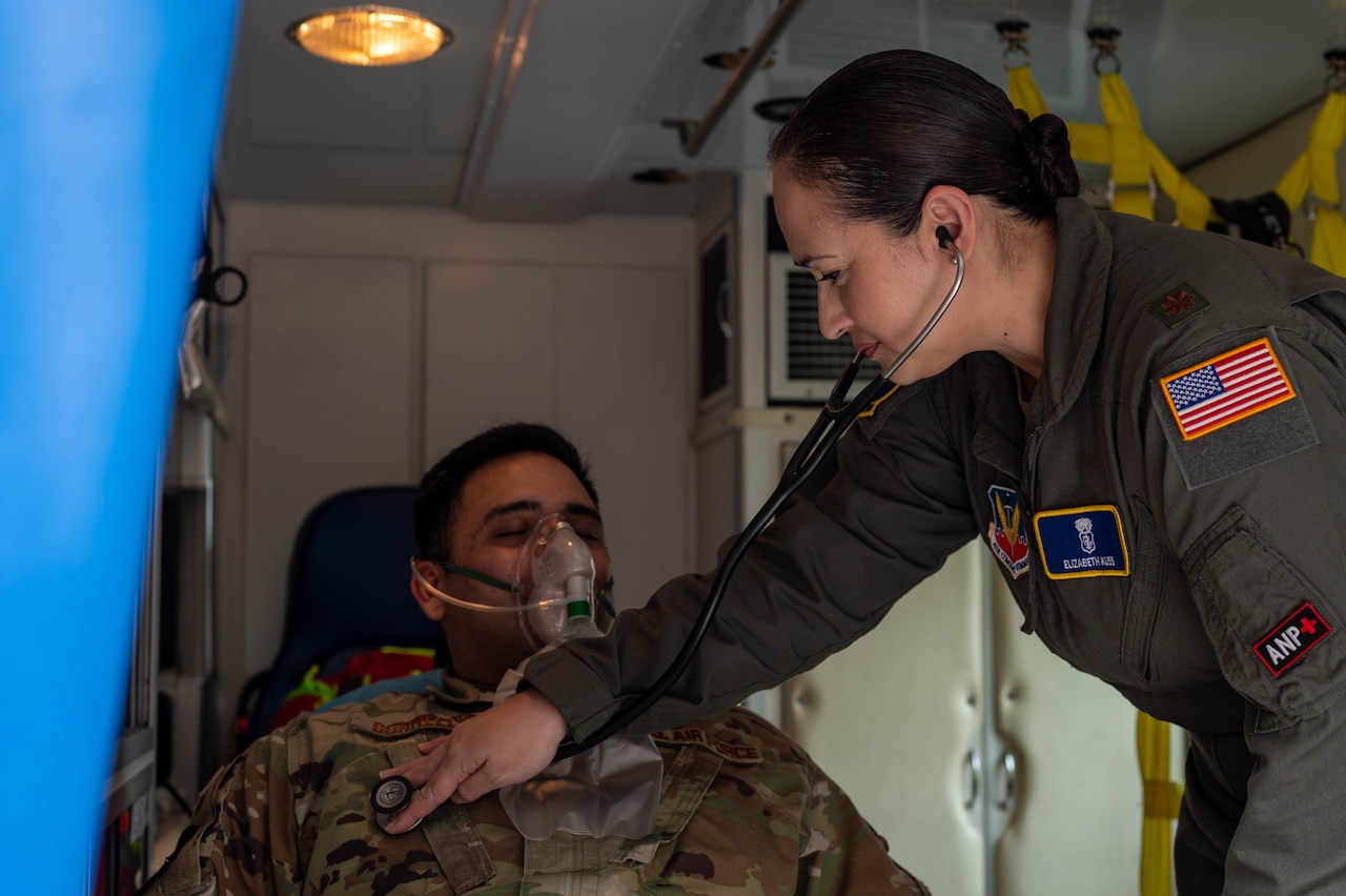 An airman in a flight suit uses a stethoscope to examine a sitting person wearing a camouflage uniform and an oxygen mask over their nose and mouth.