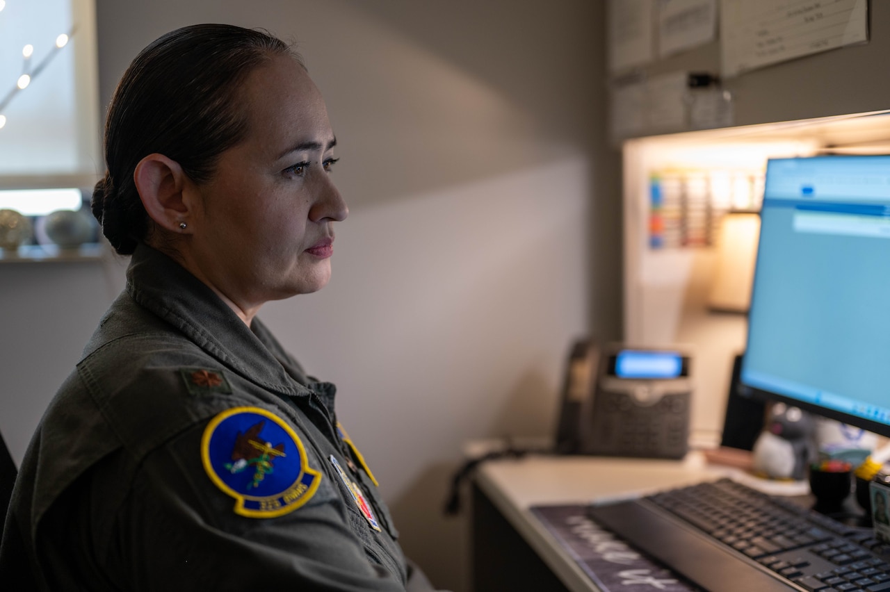 An airman in a flight suit looks at a computer screen in an indoor office setting.
