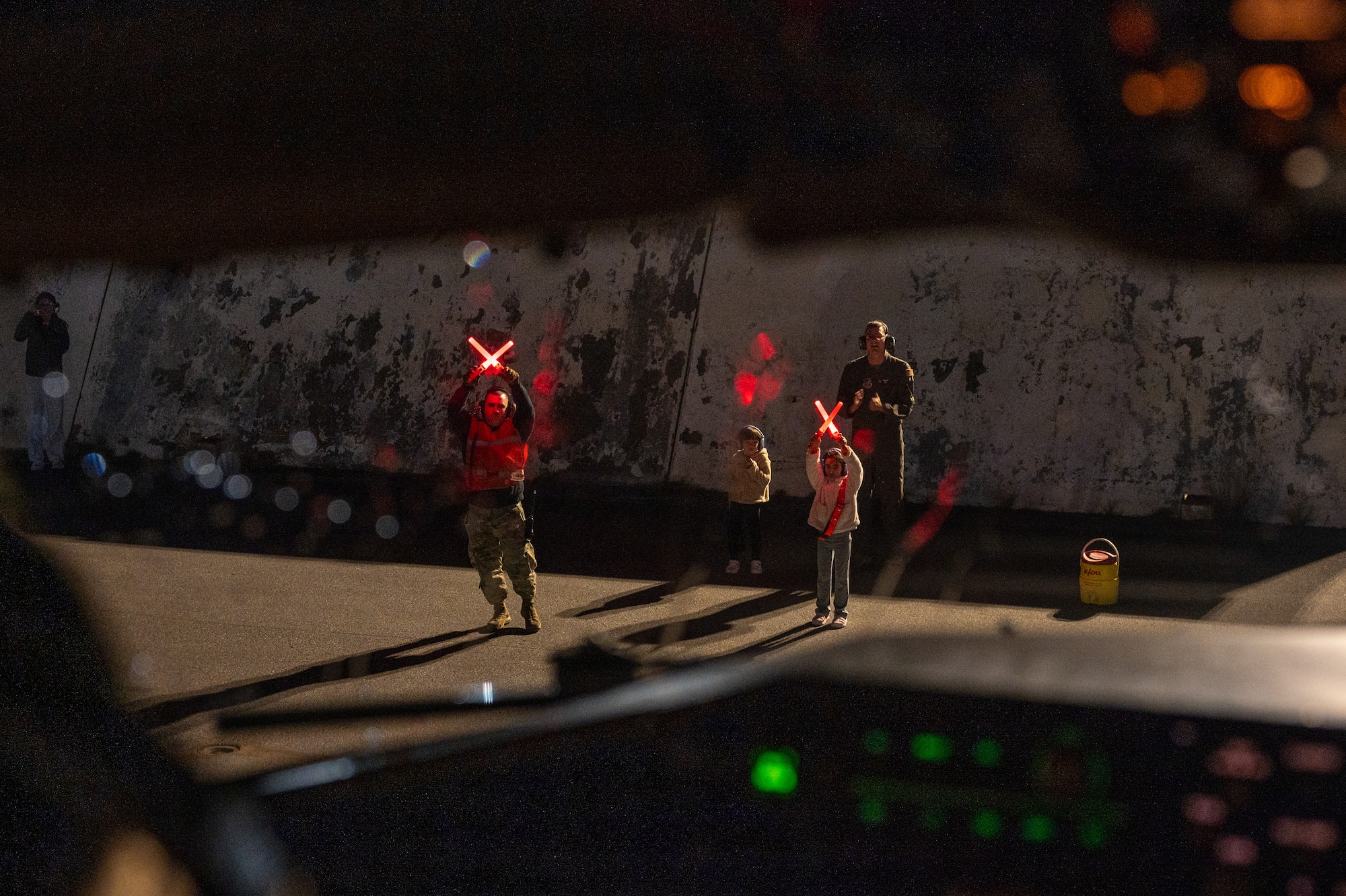 a pilots daughter and man hold marshalling lights in a crossed motion to help the pilot in the cockpit park the plant.
