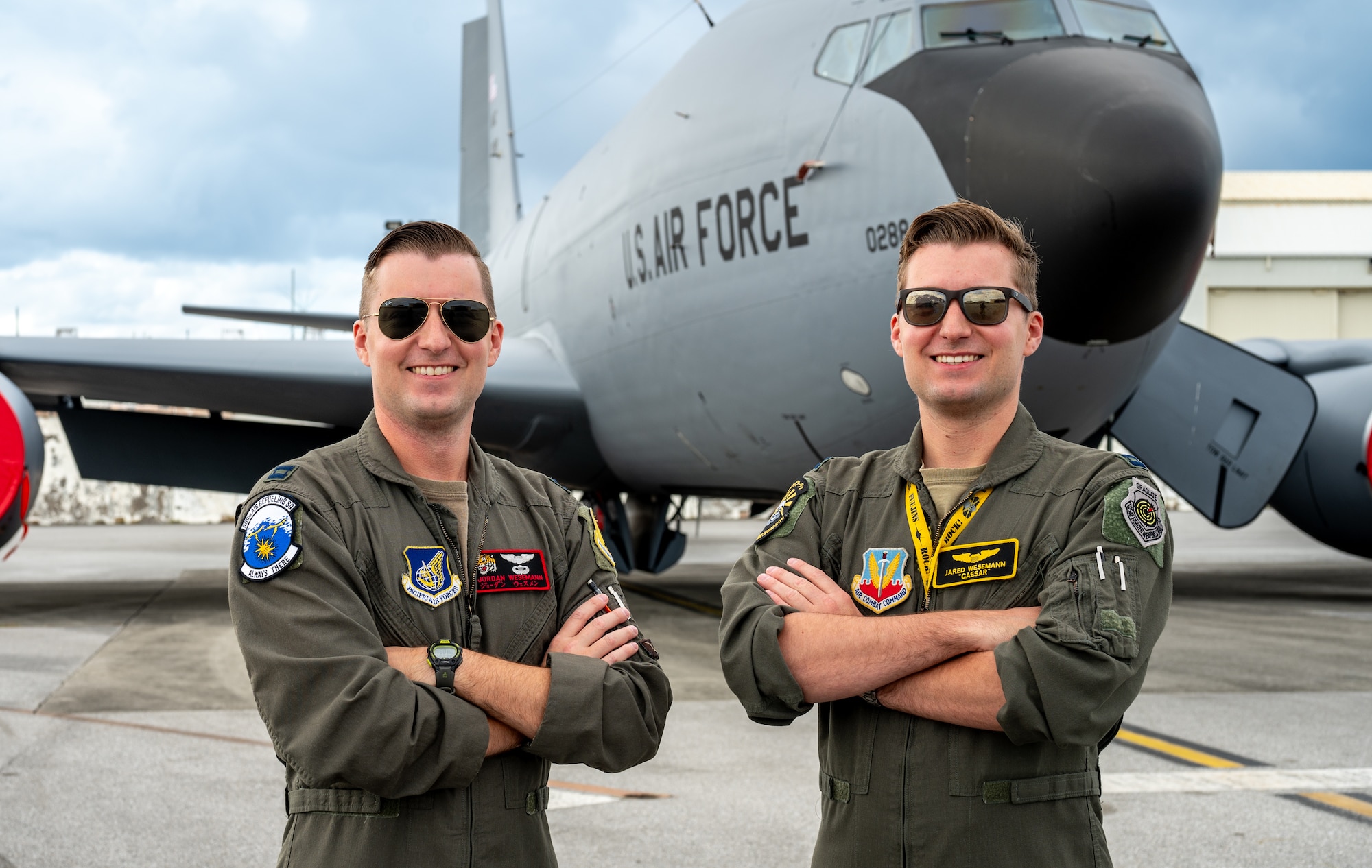 twin pilots wearing green pickle suits stand with their arms crossed smiling in front of a KC-135 Stratotanker plane