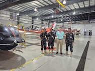 Three uniformed service members pose for a photo in a hangar with a person in civilian clothing. There are aircraft to the side and behind them.