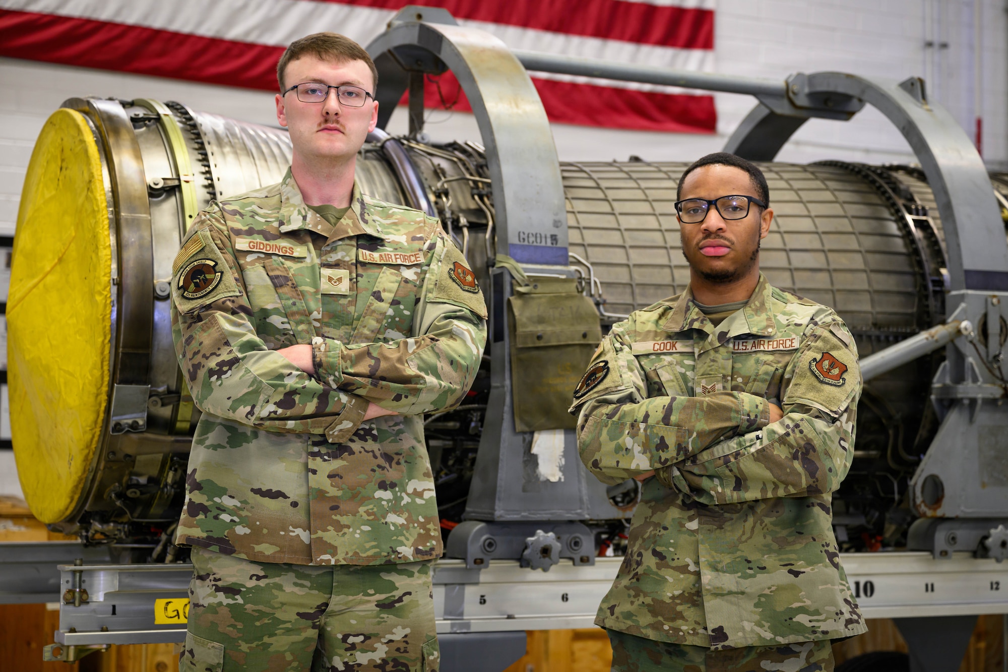 Two Airmen stand in front of an aircraft engine inside a building.