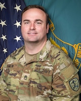 Man in U.S. Army uniform standing in front of two flags.