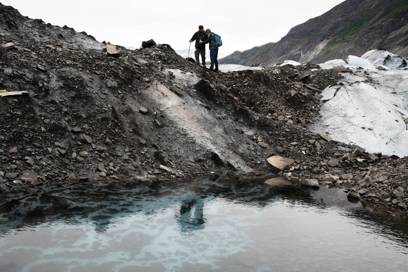 Two people stand at the top of a rocky icy hill. A pool of clear water is at the bottom of the hill.
