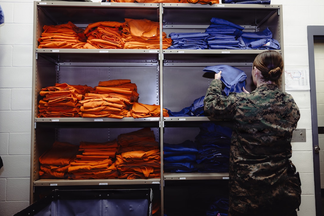 U.S. Marine Corps Lance Cpl. Mayli Motter, a facilities escort with the Marine Corps Installations East-Marine Corps Base (MCB) Camp Lejeune Regional Brig, situates prisoner uniforms on MCB Camp Lejeune, Dec. 15, 2025. Service members who have yet to undergo trial wear the blue uniform, while convicted prisoners wear the orange uniform. (U.S. Marine Corps photo by Cpl. Loriann Dauscher)