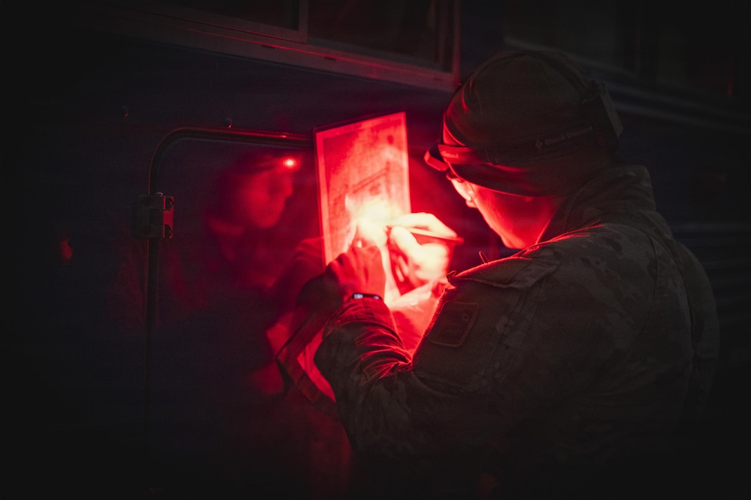 A soldier in a camouflage military uniform and a red light on their head holds a pen and looks at a navigational map at night.