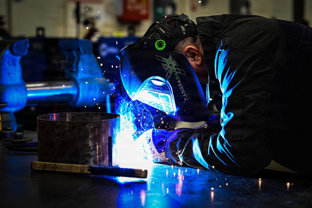 A person wearing a face shield and gloves welds a cylindrical object in a metal shop, causing blue sparks and light to emit from the object.