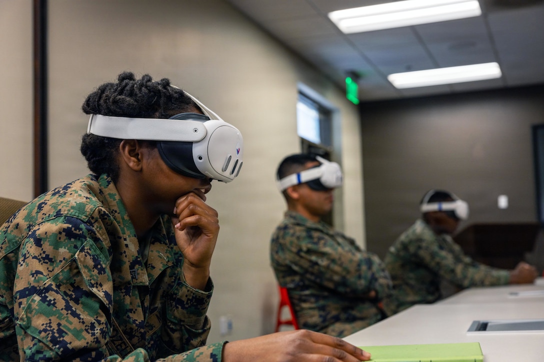 Three people wearing camouflage military uniforms and virtual reality headsets sit in a row at a table indoors.