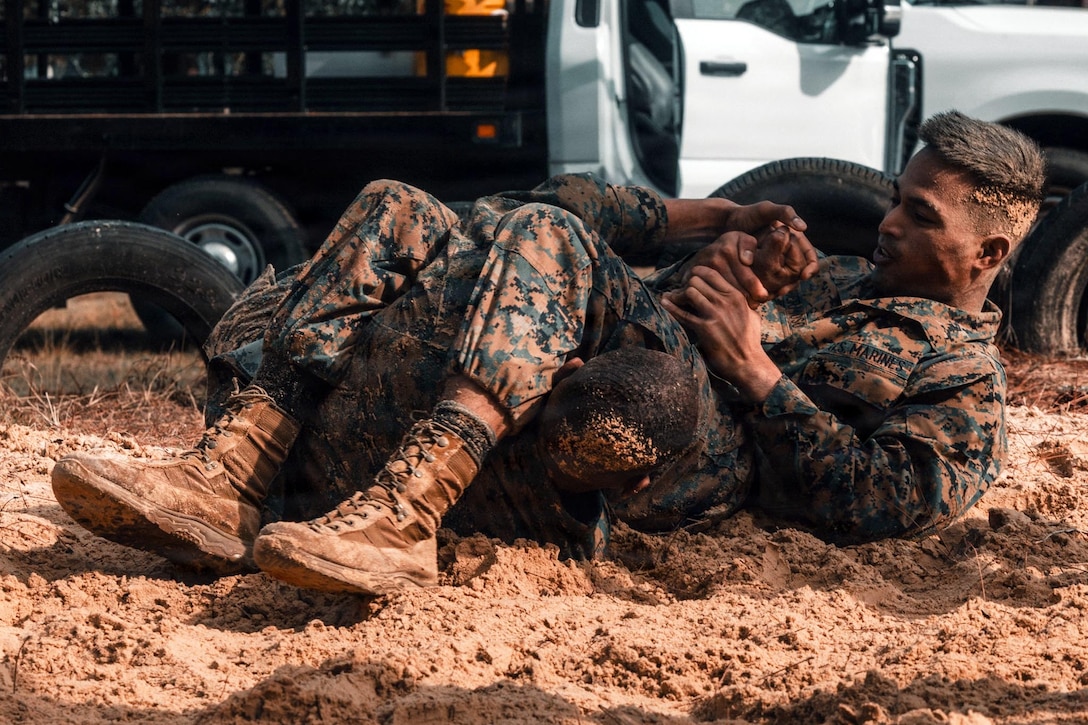 Two Marines wearing camouflage military uniforms wrestle in the sand while participating in a squad competition.