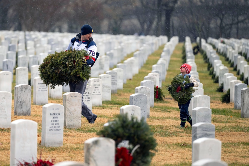 A man in cold-weather casual attire carries several wreaths looks back at a small child in similar attire, also holding a wreath, as they walk through a cemetery.