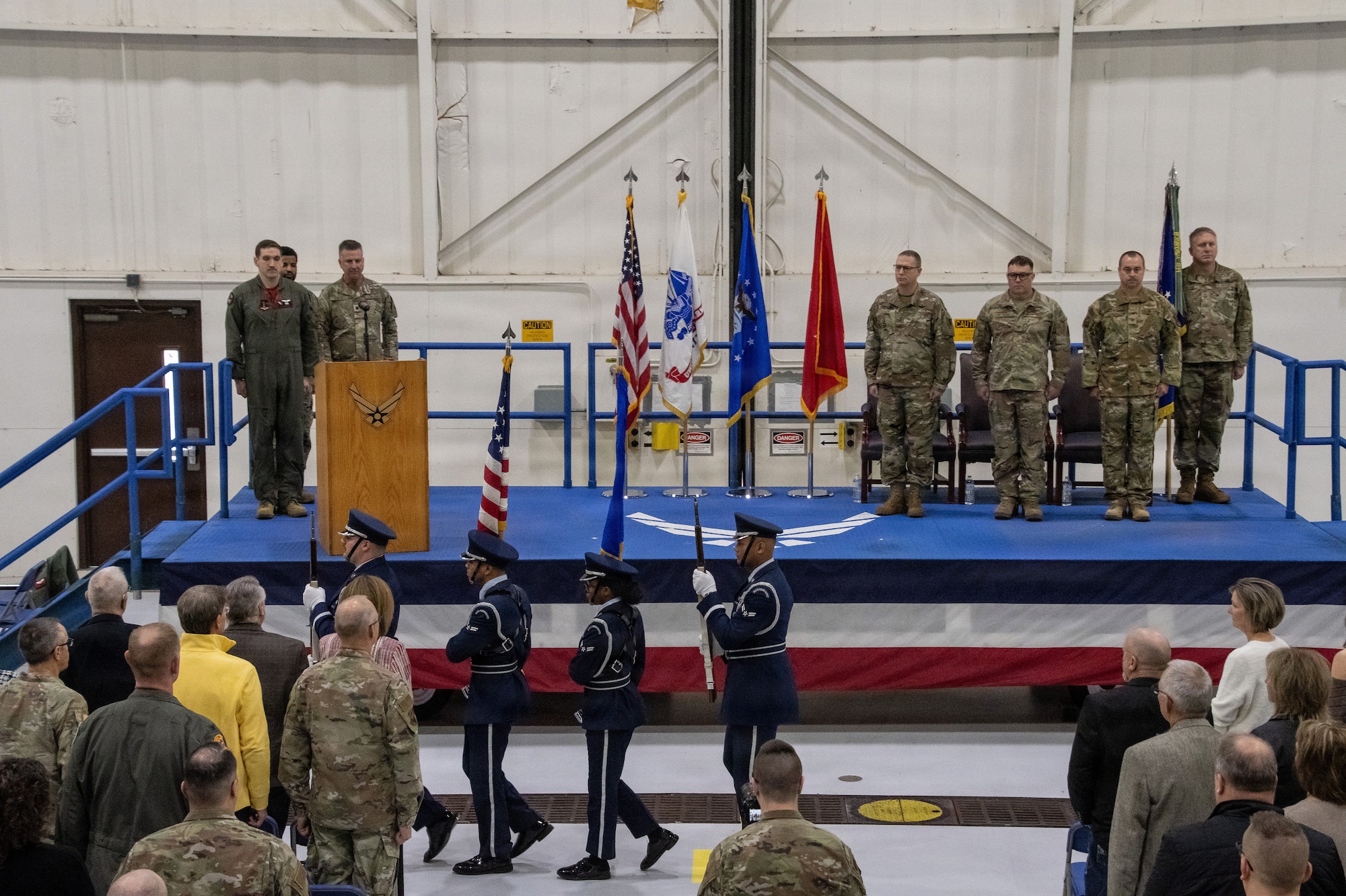 U.S. Air Force honor guard Airmen conduct a colors presentation before a ceremony. Audience members and ceremony participants stand at attention showing respect to the U.S. flag.