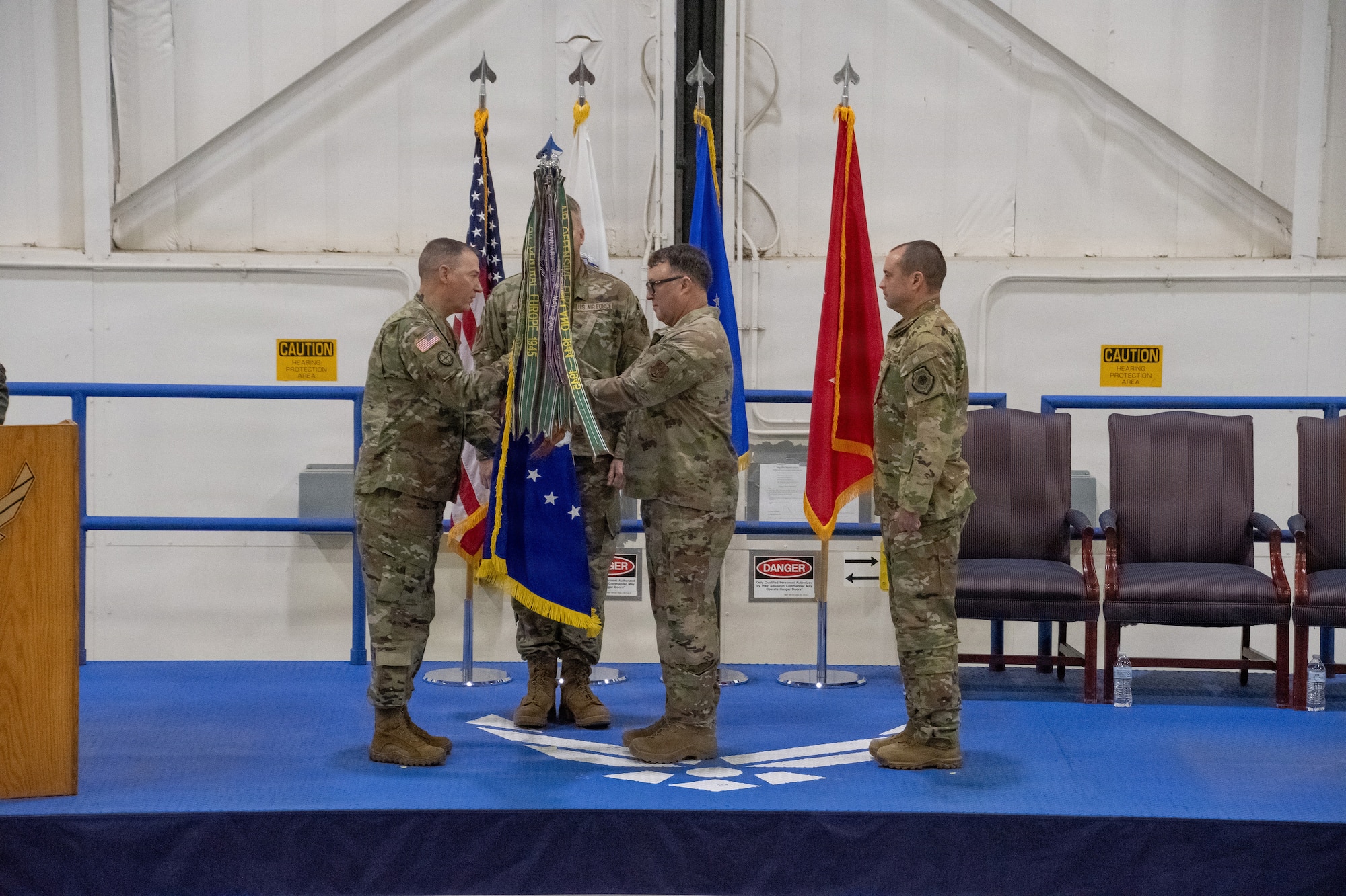 Missouri National Guard troops stand on a stage during a ceremony. Two troops exchange a guidon flag. The flag is bright blue with colorful streamers attached to the finial. The troops are wearing OCP pattern uniforms. The stage is bright blue with a large white U.S. Air Force wings logo in the center.