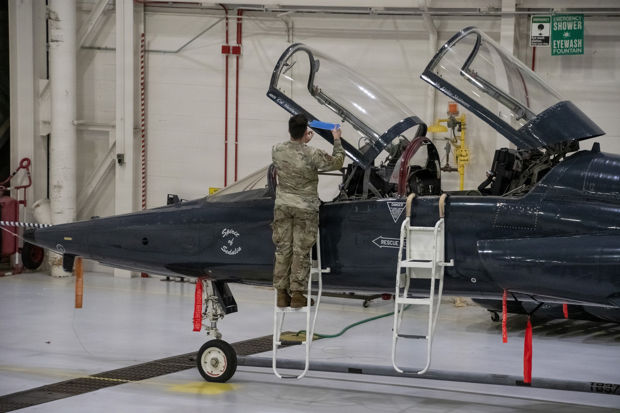 An Air National Guard Airman stands on a crew access step ladder beside a T-38 Talon trainer jet. The Airman is wearing OCP pattern uniform and removing a decal from the jet's open canopy. The jet is dark blue with white lettering and decals.