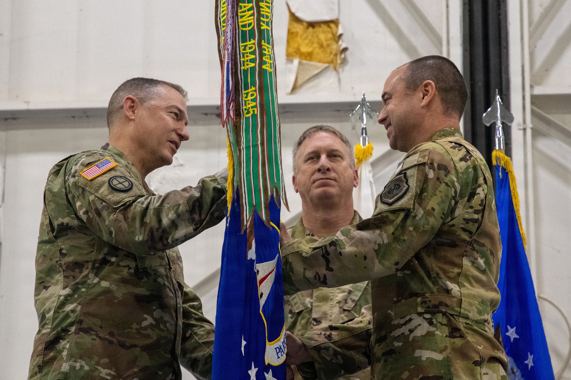 Three National Guard troops stand on a stage. They are wearing OCP pattern uniforms. Two members are exchanging a blue unit guidon flag between themselves. The flag is bright blue with colorful streamers attached to the finial.