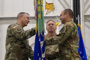 Three National Guard troops stand on a stage. They are wearing OCP pattern uniforms. Two members are exchanging a blue unit guidon flag between themselves. The flag is bright blue with colorful streamers attached to the finial.