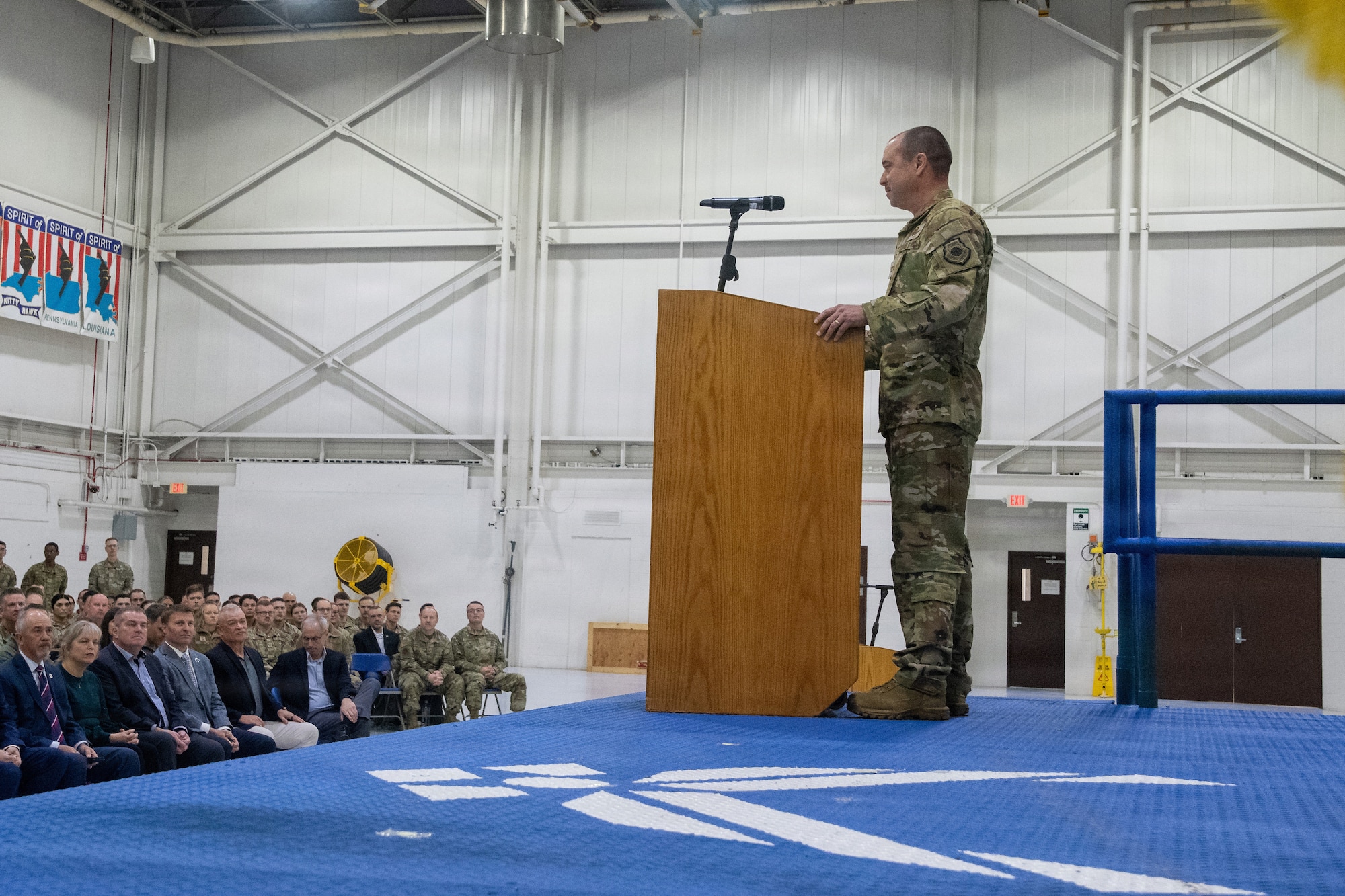 A U.S. Air National Guard colonel stands at a lectern on stage in an aircraft hangar. He is speaking to a large crowd of military members and civilians seated below the stage. A large Air Force wings logo is visible on the stage surface.