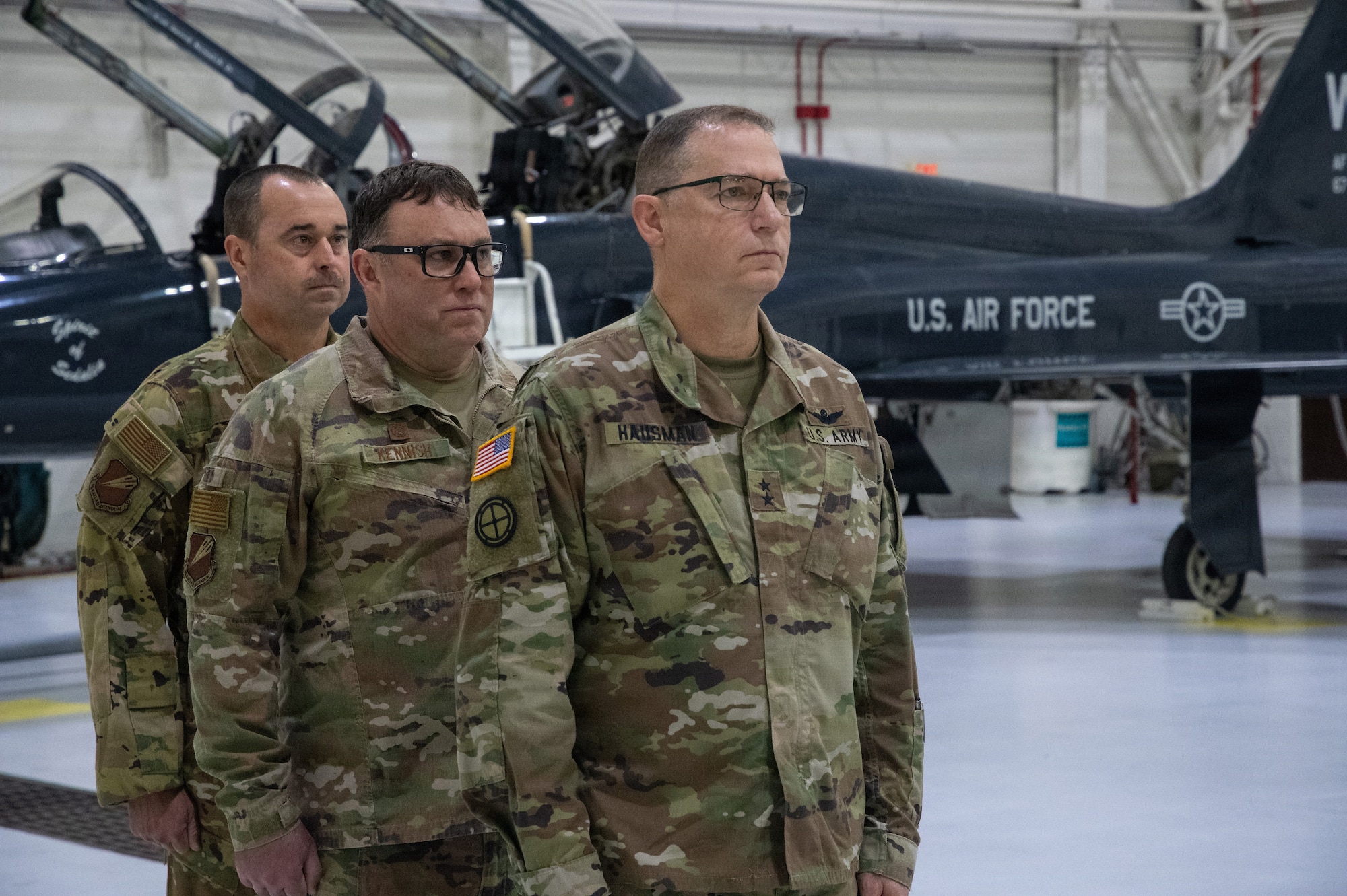 Three National Guard leaders stand at attention inside an aircraft hangar. The troops are wearing OCP pattern uniforms. A dark blue T-38 Talon trainer two-seat jet trainer is parked behind them.