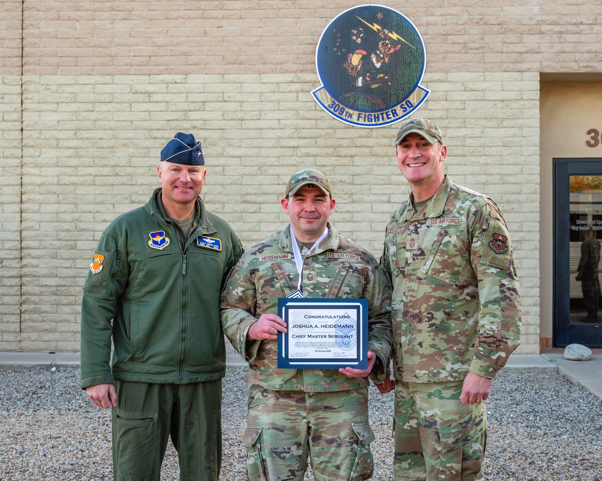 U.S. Air Force Brig. Gen. David Berkland (left), 56th Fighter Wing commander, and Chief Master Sgt. Nathan Chrestensen (right), 56th Fighter Wing command chief, pose for a photo with Senior Master Sgt. Joshua Heidemann, 56th Operations Support Squadron operations superintendent, after it was announced Heidemann was selected for promotion to Chief Master Sgt., Jan. 7, 2026, at Luke Air Force Base, Arizona.