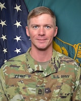 Man in U.S. Army uniform standing in front of two flags.