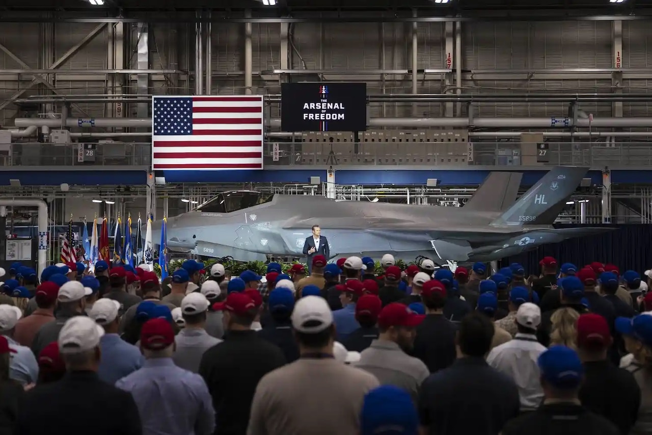 A person standing in front of a fighter jet parked indoors addresses a crowd of people, with an American flag and a sign reading "The Arsenal of Freedom" in the background.