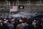 A person standing in front of a fighter jet parked indoors addresses a crowd of people, with an American flag and a sign reading "The Arsenal of Freedom" in the background.