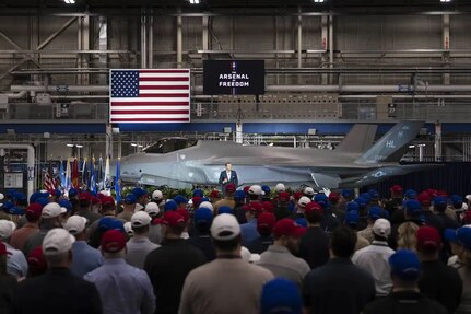 A person standing in front of a fighter jet parked indoors addresses a crowd of people, with an American flag and a sign reading "The Arsenal of Freedom" in the background.
