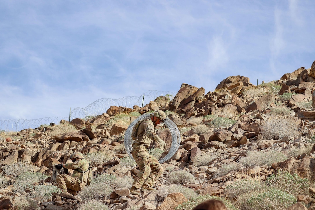 One soldier sits as another walks with concertina wire on a mountain under a cloudy, blue sky.