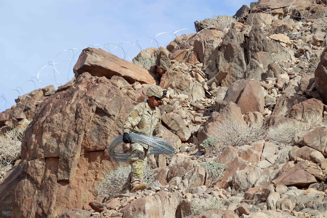 A soldier in protective gear carries concertina wire on a mountain under a cloudy, blue sky.