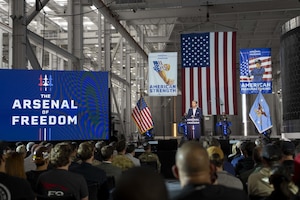 The secretary of war stands in front a large American flag with speaking at a podium to an audience with a monitor to the left.