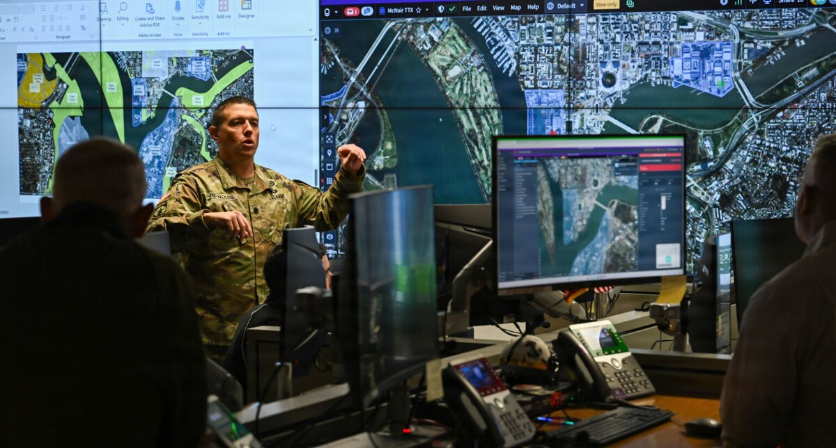 A man in a camouflage military uniform stands in front of a wall of screens with maps on them as he talks to people in the foreground.