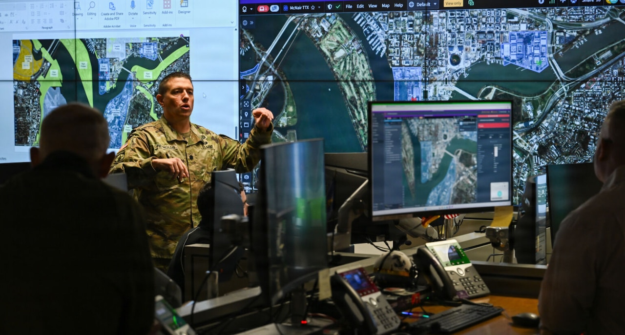 A man in a camouflage military uniform stands in front of a wall of screens with maps on them as he talks to people in the foreground.