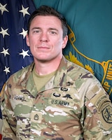 Man in U.S. Army uniform standing in front of two flags.