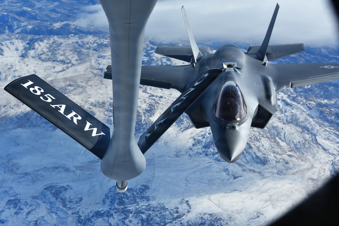 A refueling boom hovers near a military jet high above snow-capped mountains.