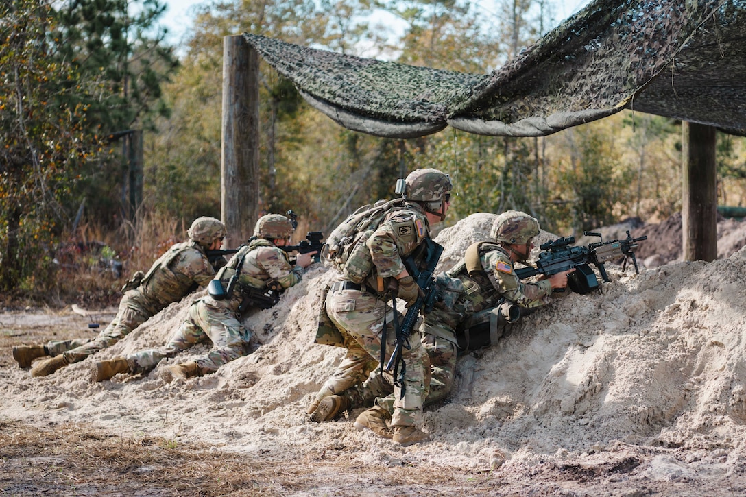 Soldiers in tactical gear rest on mounds of dirt while aiming weapons under a net in a wooded area.