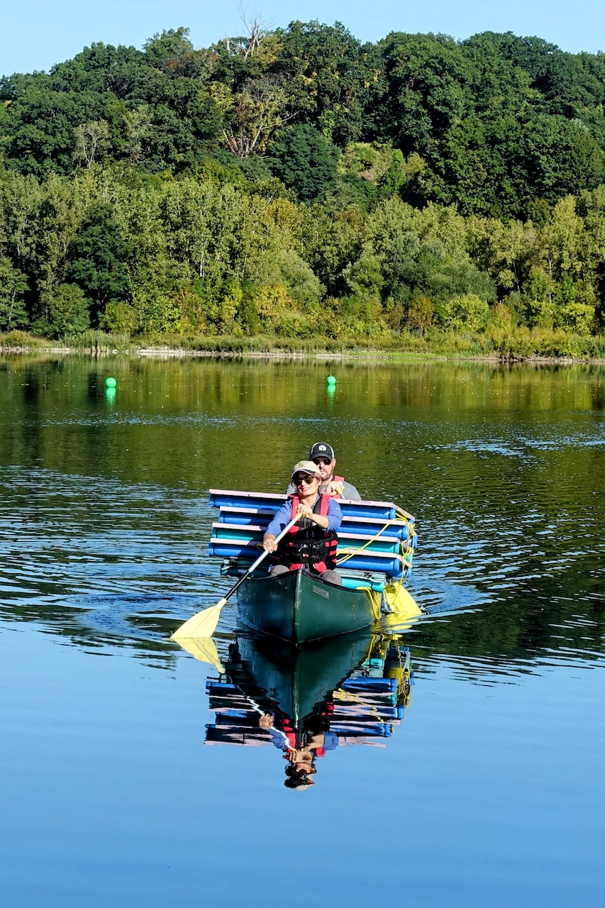 Two people cross a body of water in a boat while carrying large square metal panels.