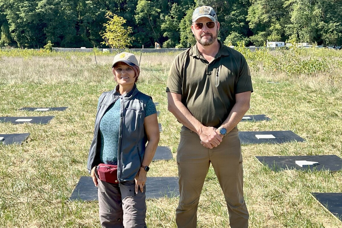 A woman and a man dressed in civilian attire stand in front of panels that are on the ground in a field, with trees in the background.