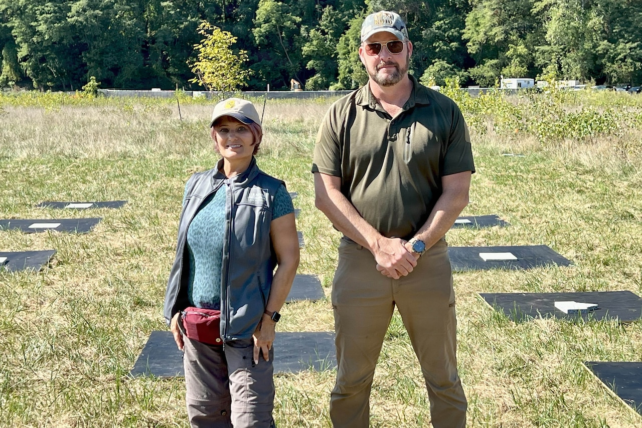 A woman and a man dressed in civilian attire stand in front of panels that are on the ground in a field, with trees in the background.