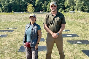 A woman and a man dressed in civilian attire stand in front of panels that are on the ground in a field, with trees in the background.