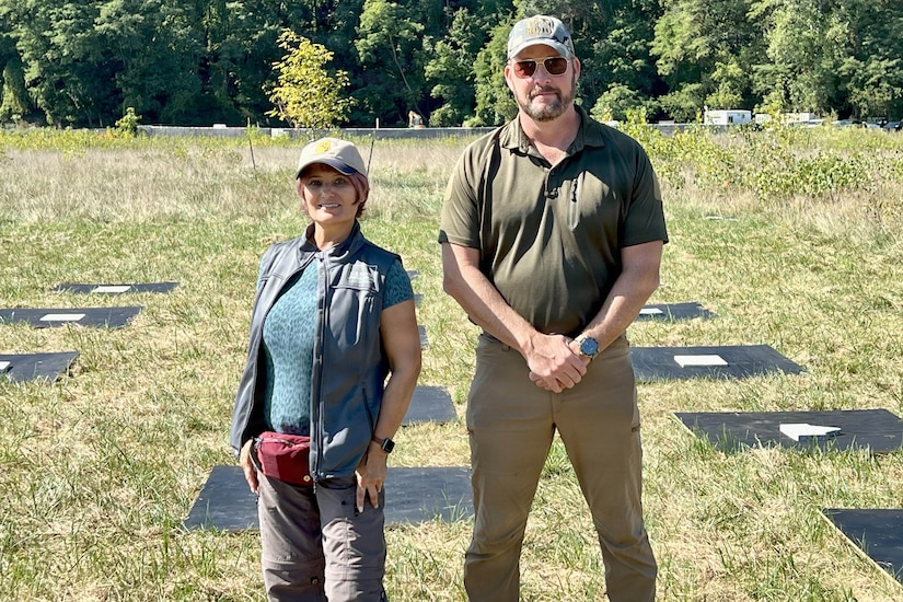 A woman and a man dressed in civilian attire stand in front of panels that are on the ground in a field, with trees in the background.