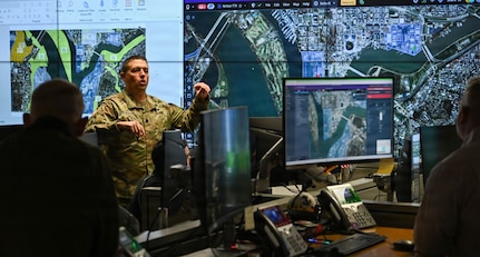 A man in a camouflage military uniform stands in front of a wall of screens with maps on them as he talks to people in the foreground.