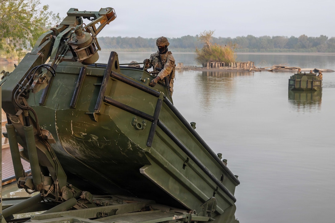 A soldier operates a bridge-building boat in a body of water, with another boat cruising in the background during daytime.