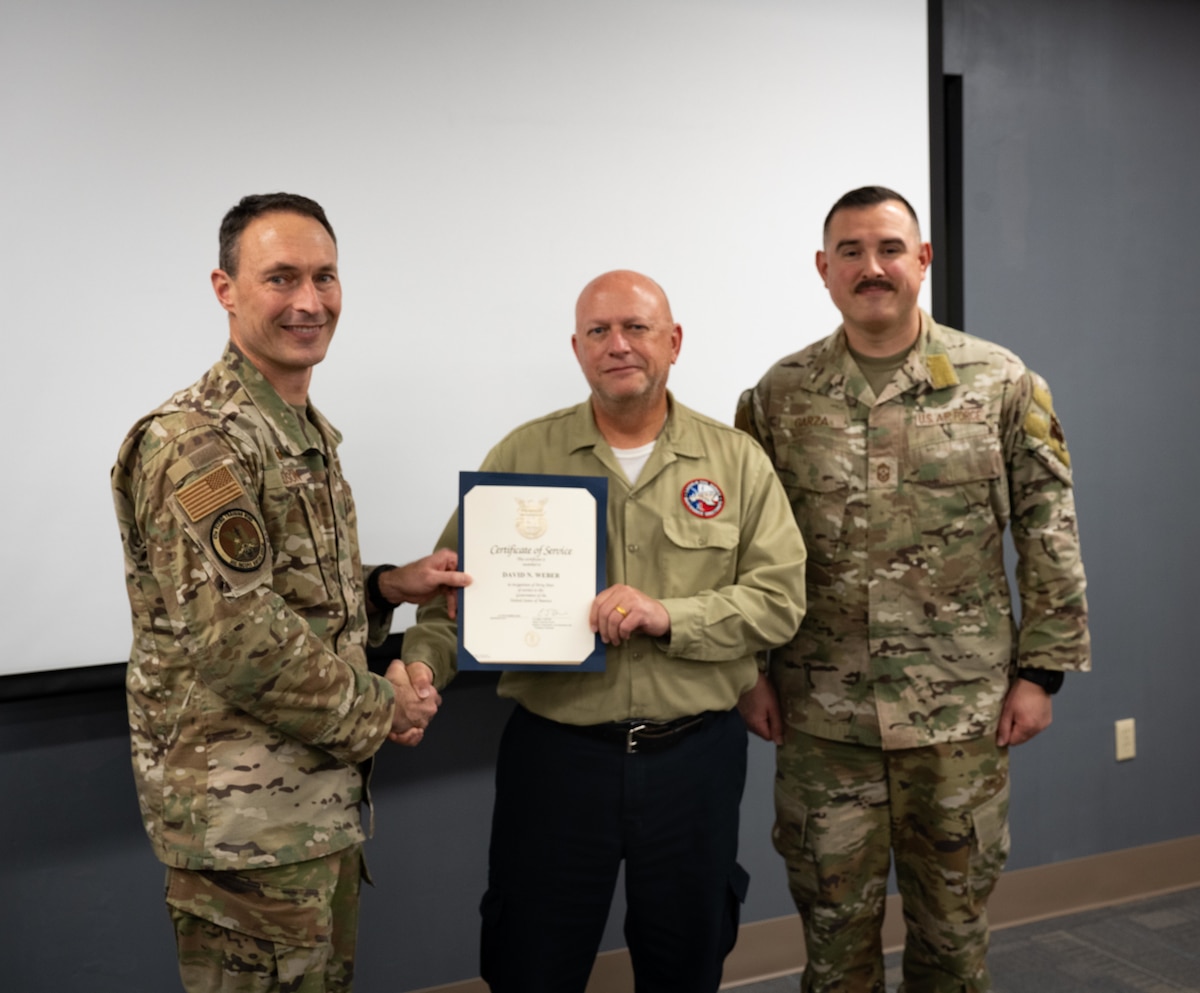 U.S. Air Force Col. Tyler Ellison, 47th Flying Training Wing commander, and U.S. Air Force Chief Master Sgt. Erik Garza, 47th Flying Training Wing command chief, presents a certificate of service to David Weber (center), Quality Assurance Environmental and Safety Programs Manager, during a recognition ceremony at Laughlin Air Force Base, Texas, Dec. 4, 2025, celebrating Weber’s more than 40 years of federal service. Chief Master Sgt. Erik Garza (right), 47th Maintenance Directorate senior enlisted leader, attends the presentation. (U.S. Air Force photo by Airman 1st Class Harrison Sullivan)