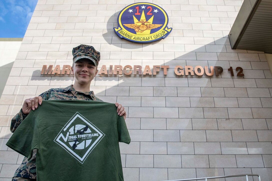 U.S. Marine Corps Lance Cpl. Cecilia Campbell, a combat videographer with Marine Aircraft Group 12, 1st Marine Aircraft Wing, presents a shirt given to her during a visual storytelling workshop while posing for a picture at Marine Corps Air Station Iwakuni, Japan, Dec. 15, 2025. During participation in the Marine Corps Instillations Pacific hosted course, she received first place in the photo story and b-roll package portions. Campbell is a native of Virginia. (U.S. Marine Corps photo by Lance Cpl. David Getz)