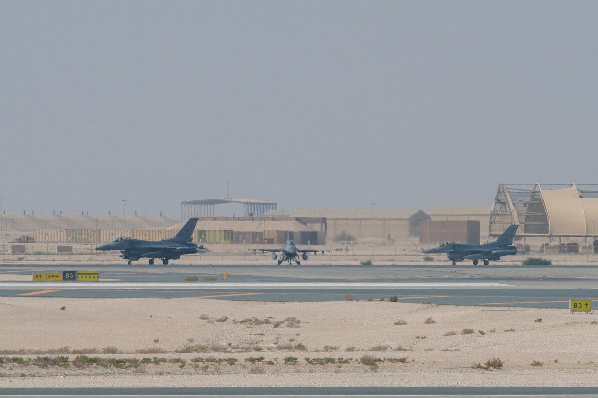Three military fighter jets taxi on a tarmac in the desert. There are aircraft facilities and shipping containers in the background.