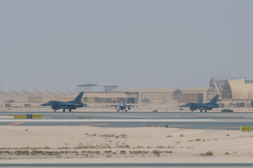 Three military fighter jets taxi on a tarmac in the desert. There are aircraft facilities and shipping containers in the background.