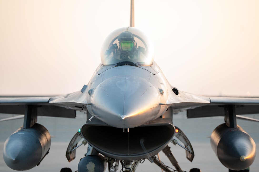 A close-up view of a service member in a flight suit and helmet sitting in the cockpit of a military fighter jet on a tarmac.