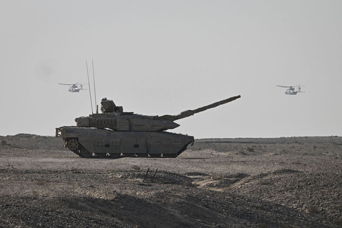 A large military tank points its cannon in the air while sitting in the desert. Two military helicopters are flying in the background.