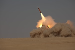 A rocket launches from a stand, with flames shooting out the back and a cloud of dust coming off the ground.