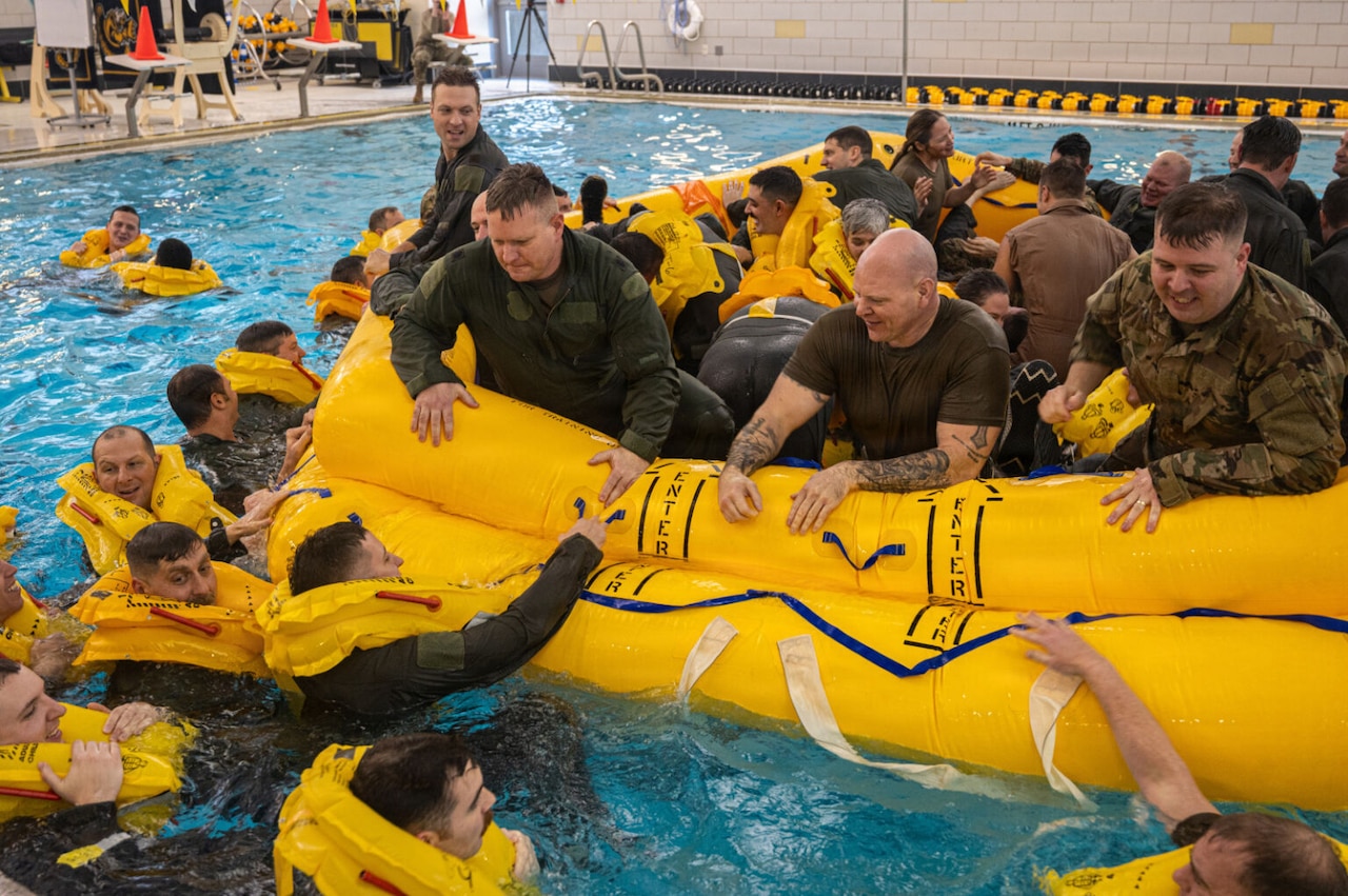 A group of military personnel sit inside a life raft in an indoor pool. Other people in military uniform swim outside the life raft.