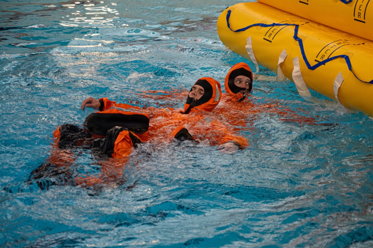 Two people in orange water survival suits, swim toward a life raft in a large indoor pool.